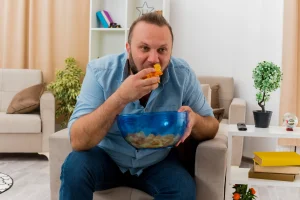 joyful adult slavic man sits armchair holding eating bowl chips inside living room