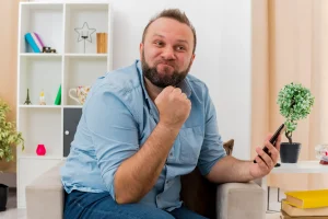 annoyed adult slavic man sits armchair keeping fist holding phone inside living room